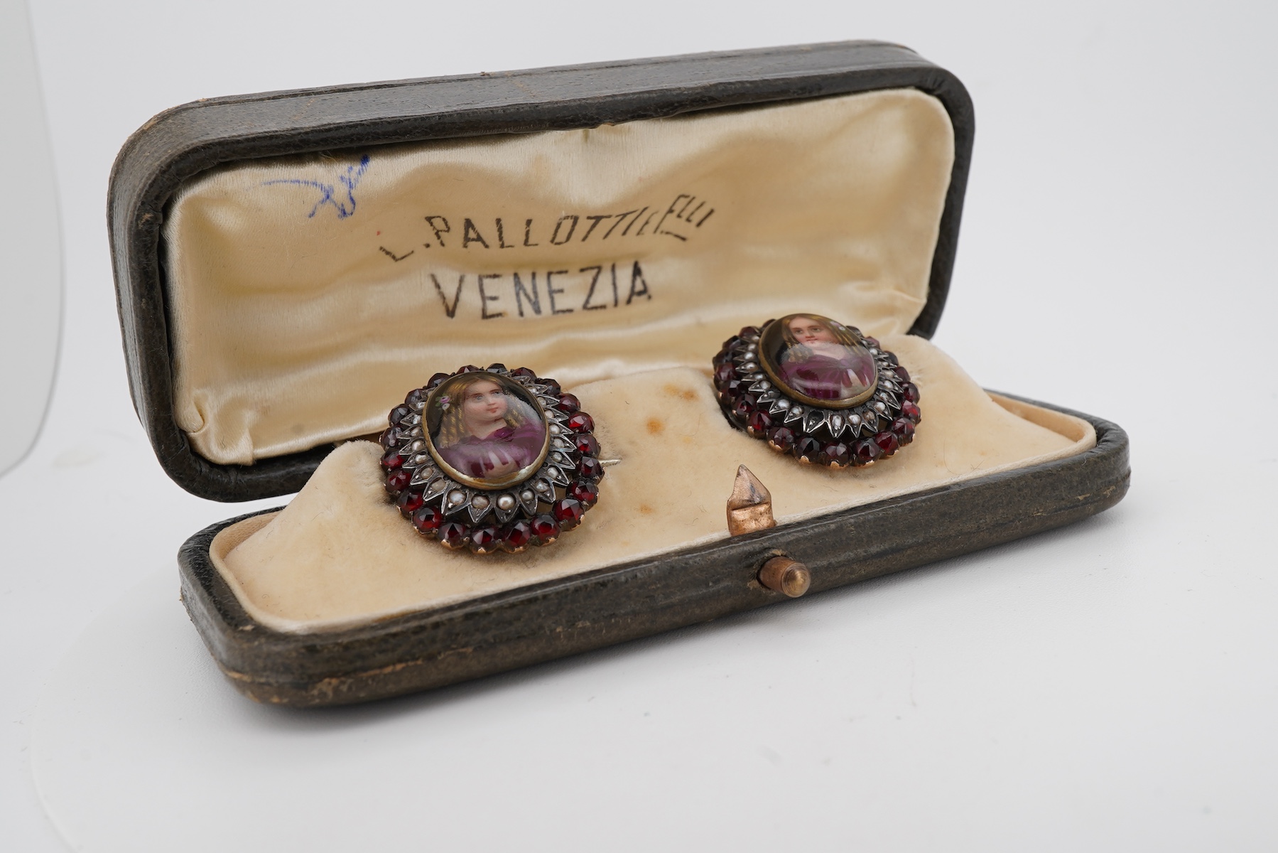 A pair of early 20th century yellow metal mounted brooches, with central oval panel painted with the bust of a lady and bordered by concentric rows of seed pearls and garnets (adapted), 24mm by 20mm.